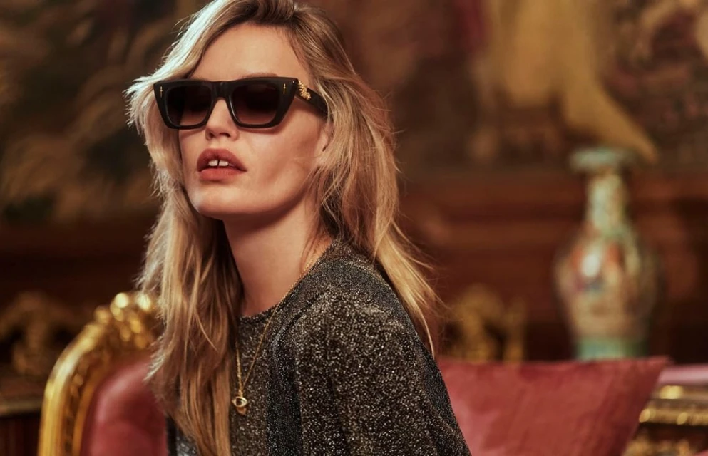 Adult female wearing designer sunglasses and a shimmering black blouse, gazing at the camera in a luxurious indoor setting.