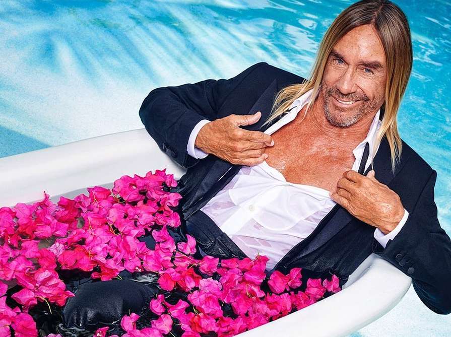 Iggy Pop reclines in a bathtub filled with pink bougainvillea flowers, wearing a suit, open shirt, smiling against an aqua pool background.