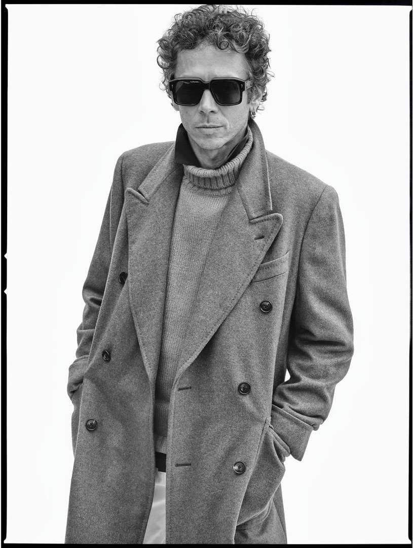 Monochrome portrait of a man with curly hair, wearing sunglasses, a gray coat, and a turtleneck, posing against a white background.
