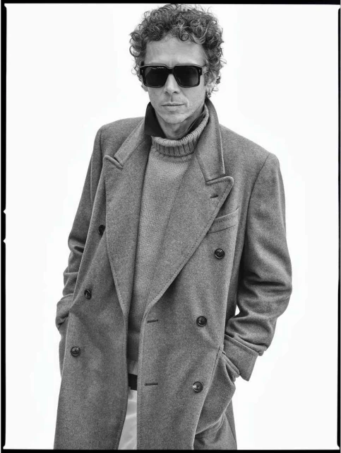 Monochrome portrait of a man with curly hair, wearing sunglasses, a gray coat, and a turtleneck, posing against a white background.