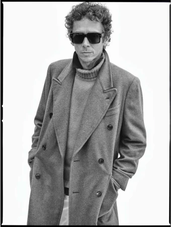 Monochrome portrait of a man with curly hair, wearing sunglasses, a gray coat, and a turtleneck, posing against a white background.