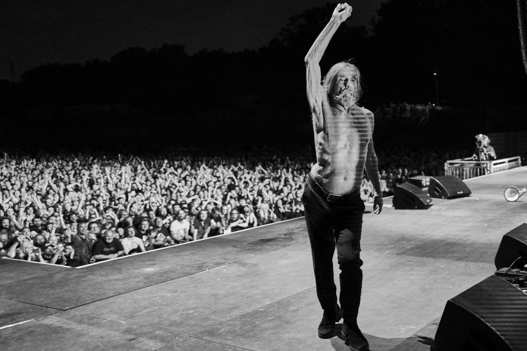 Iggy Pop performs shirtless on stage with fist raised for a large concert audience, showcasing a monochrome concert photography style.