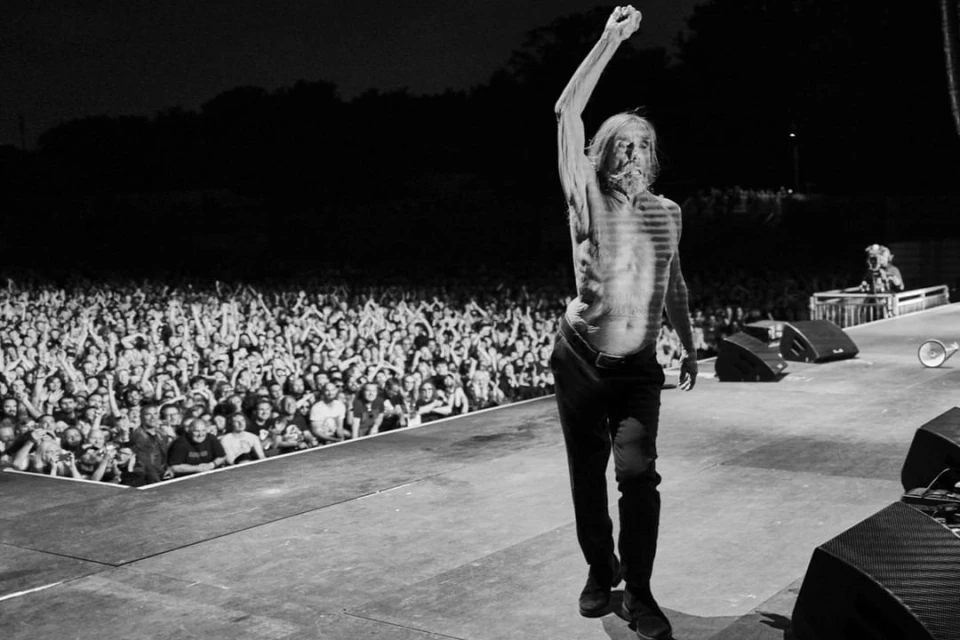 Iggy Pop performs shirtless on stage with fist raised for a large concert audience, showcasing a monochrome concert photography style.