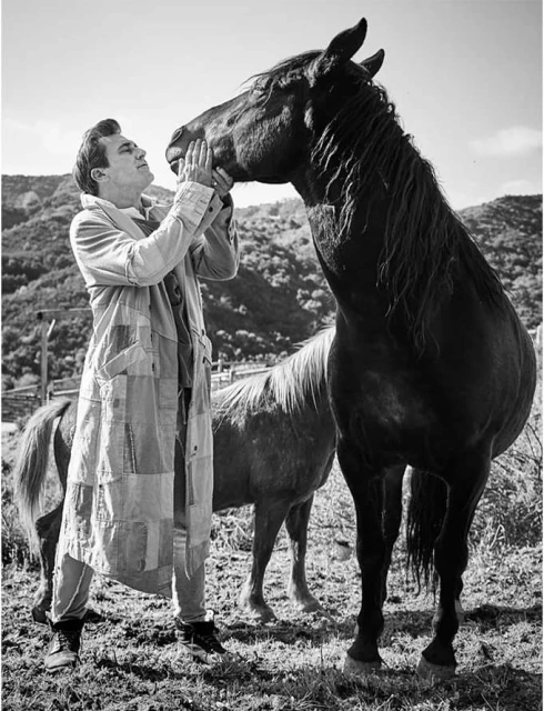 Monochrome portrait of an adult man in a patchwork coat touching noses with a dark horse in a rural, grassy, mountainous setting.