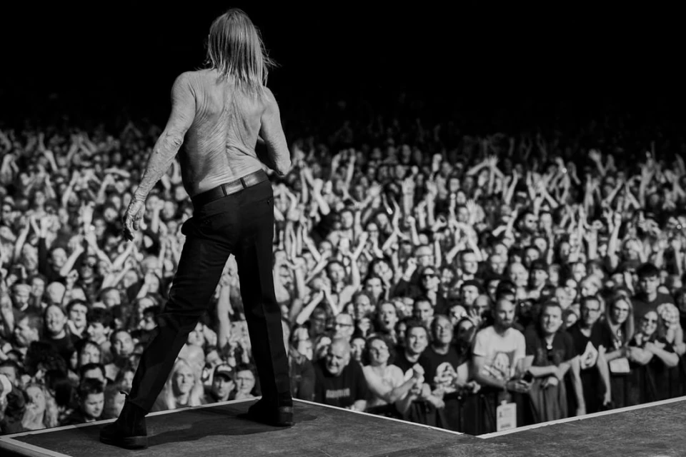 Back view of bare-chested male performer on stage facing a large audience in a dark, concert-style setting, black and white.