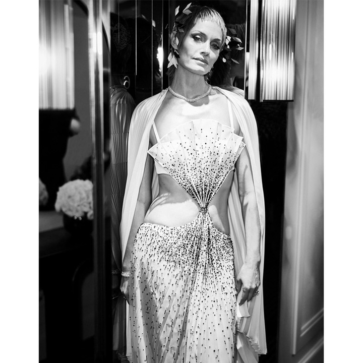 Black and white portrait of a woman in a heavily embellished gown and cape standing indoors