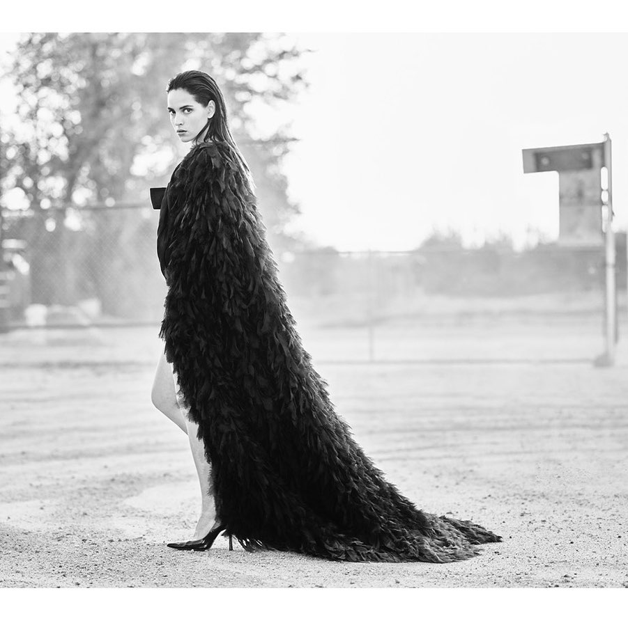 Monochrome portrait of a woman in a feathered gown, striking a pose on a sand-covered ground with trees and an urban structure in the backdrop.