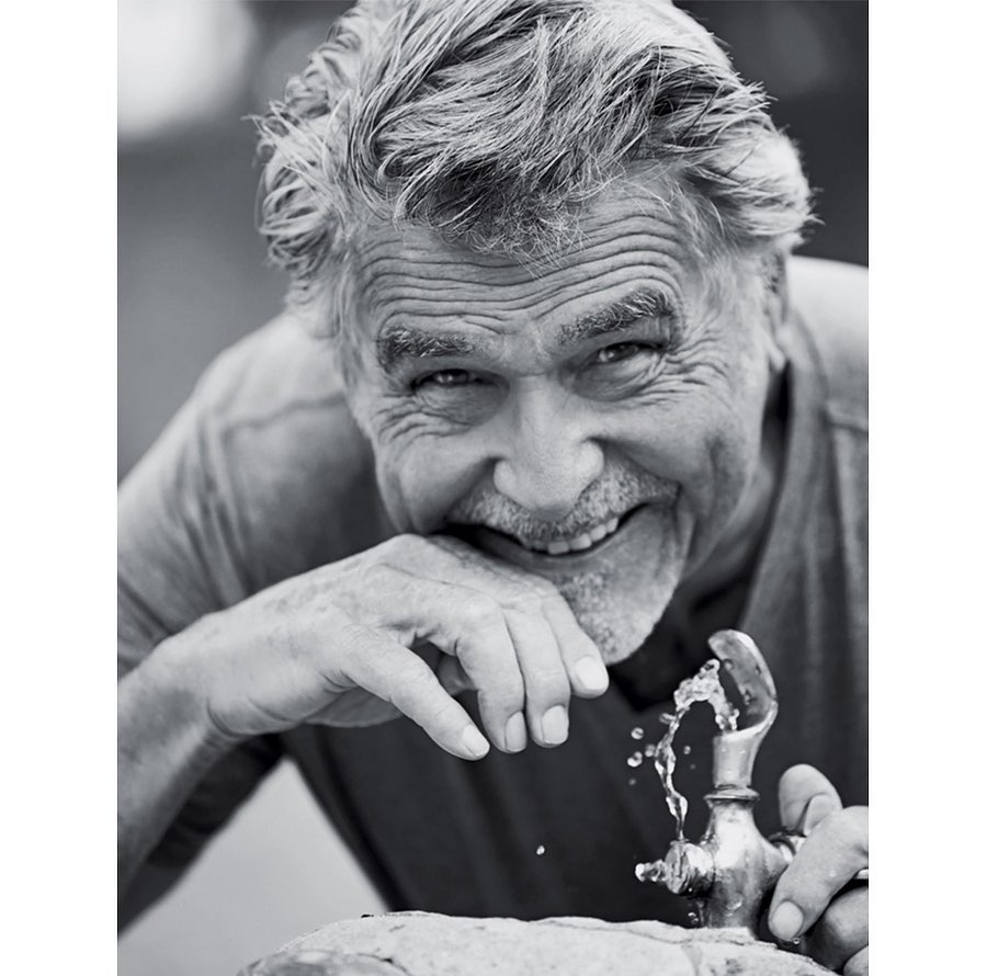 Black and white portrait of an older man with gray hair smiling and drinking water from a metal outdoor water fountain.