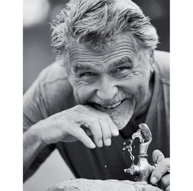 Black and white portrait of an older man with gray hair smiling and drinking water from a metal outdoor water fountain.