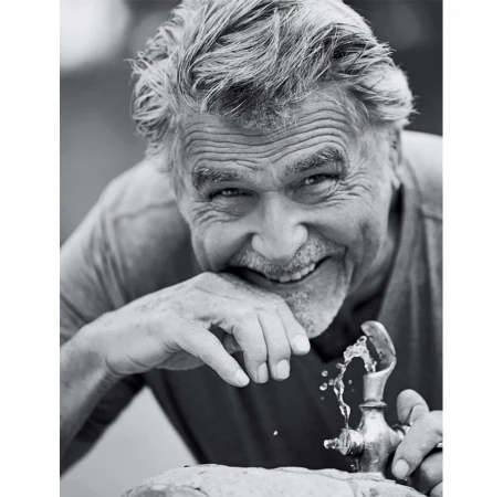 Black and white portrait of an older man with gray hair smiling and drinking water from a metal outdoor water fountain.