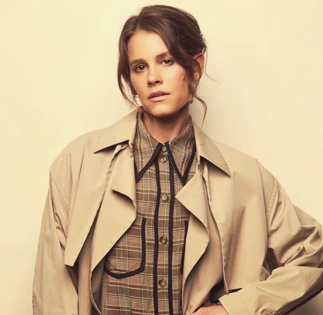 Adult woman posing in a studio shot with a plaid shirt and a trench coat. The model is looking at the camera with a neutral expression.