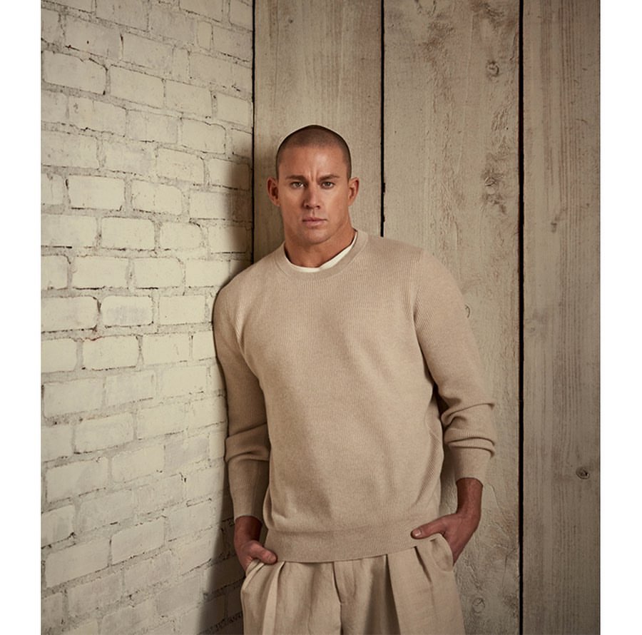 Studio shot of a man in a beige sweater and pants leaning against a white brick wall, with a weathered wood wall in the background.
