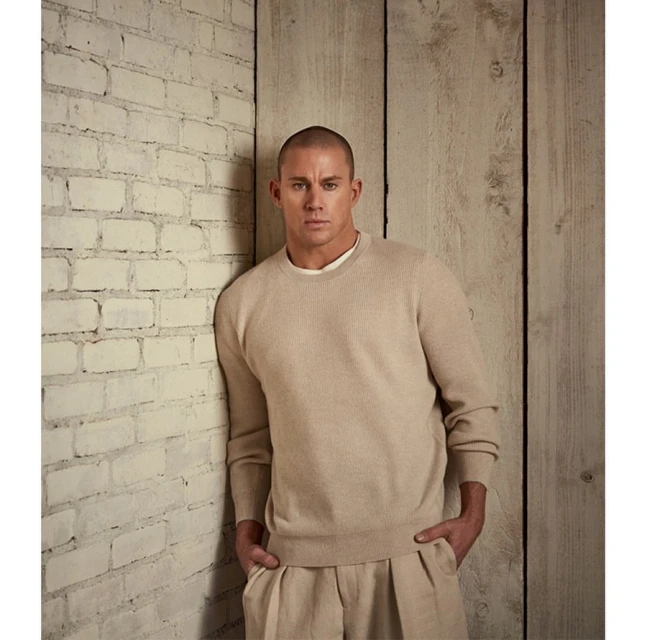 Studio shot of a man in a beige sweater and pants leaning against a white brick wall, with a weathered wood wall in the background.