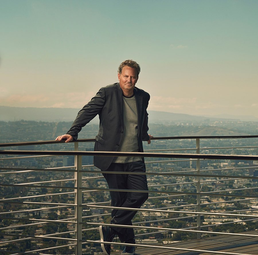 Matthew Perry leaning on a balcony railing with a city skyline visible in the background, photographed by Brian Bowen Smith.