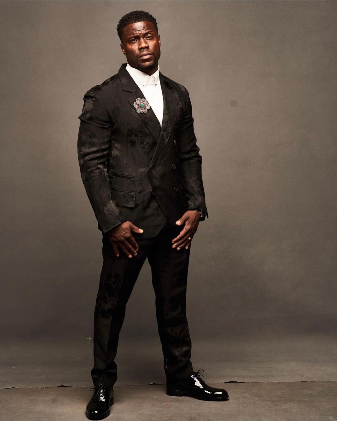 Full-length portrait of a smiling African American man in a dark patterned suit and white bow tie, standing against a gray background.