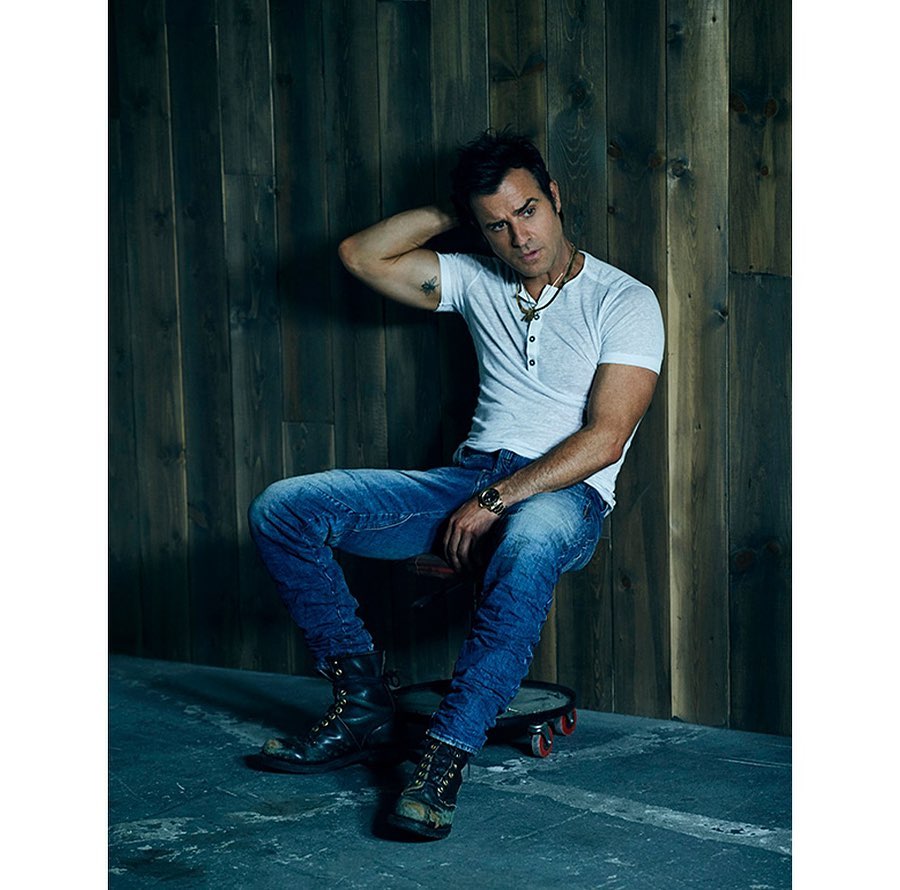 Portrait of a man in a white shirt and jeans sitting on a wheeled stool in front of a dark wood wall, looking cool and casual.