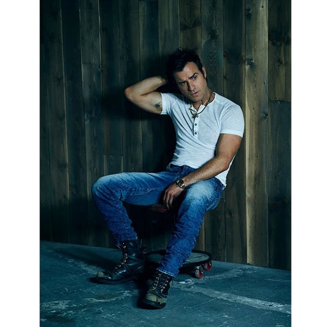 Portrait of a man in a white shirt and jeans sitting on a wheeled stool in front of a dark wood wall, looking cool and casual.