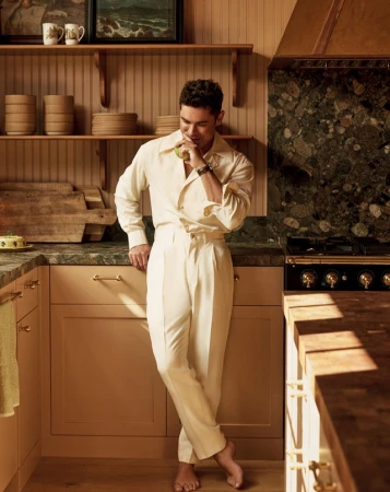 Man in off-white shirt and trousers stands in a kitchen with wooden cabinets and shelves, holding a green apple in his hand.