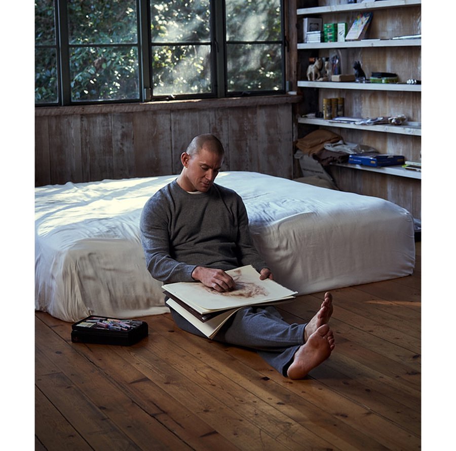 Adult man sits on a wood floor with an open sketchbook in his lap near a bed, lit by natural light in a rustic indoor setting.