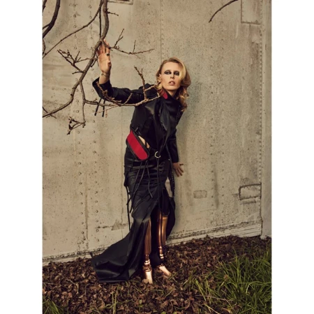 Woman in a black leather jacket and dress poses against a textured wall with bare tree branches, showing edgy fashion style.