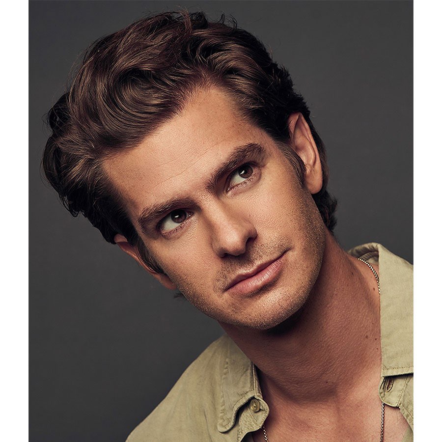 Portrait of Andrew Garfield with brown wavy hair wearing a tan shirt against a dark grey background