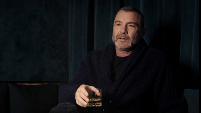 Adult male with salt-and-pepper beard sits indoors holding a glass of amber liquid, wearing a dark cardigan against a dark teal background.