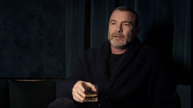 Adult male with salt-and-pepper beard sits indoors holding a glass of amber liquid, wearing a dark cardigan against a dark teal background.