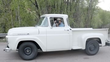 Classic white Ford F-100 pickup truck on an asphalt road, surrounded by lush green trees and overcast skies. Man visible in the cab.