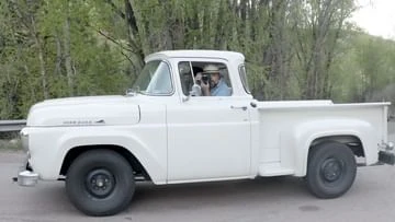 Classic white Ford F-100 pickup truck on an asphalt road, surrounded by lush green trees and overcast skies. Man visible in the cab.