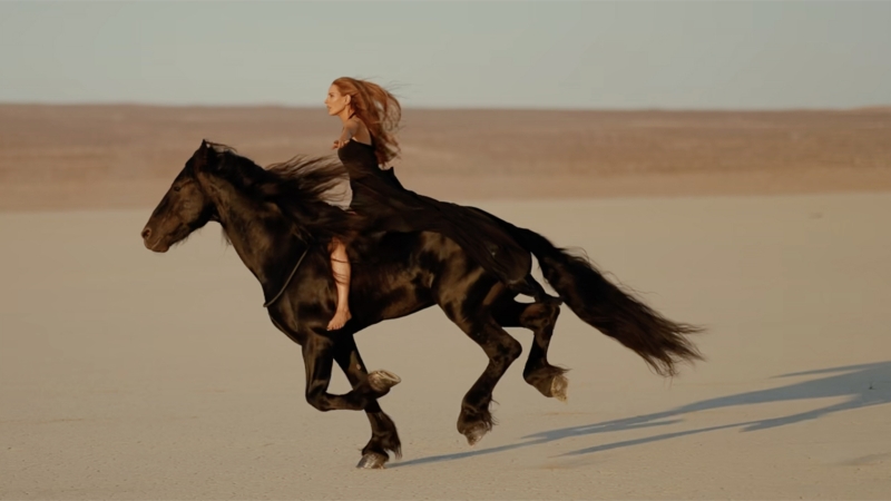 A black horse and a woman with long hair ride across a desert landscape with a tan horizon under a pale blue sky in outdoor shot.