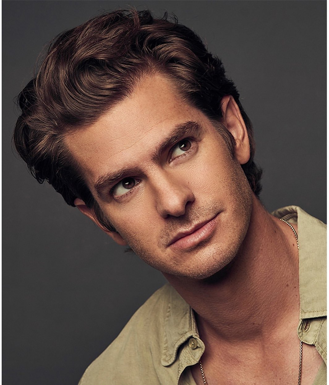 Headshot of a man with brown wavy hair, light brown skin, and a light-colored shirt, posing against a solid grey background.