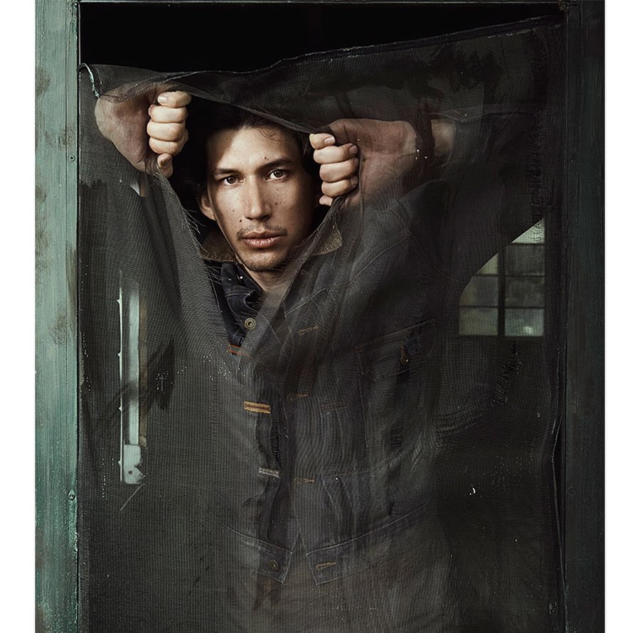 A man stands behind a mesh screen door, wearing a denim jacket, with a neutral expression in a dimly lit, indoor environment.