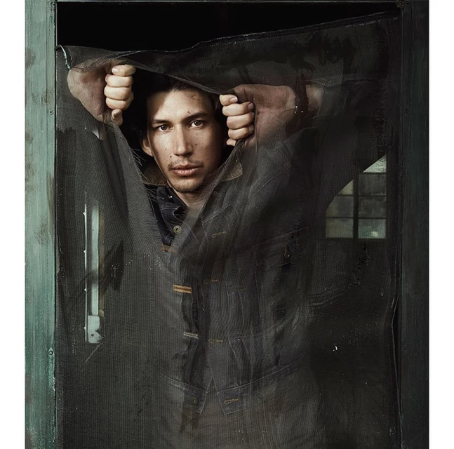 A man stands behind a mesh screen door, wearing a denim jacket, with a neutral expression in a dimly lit, indoor environment.