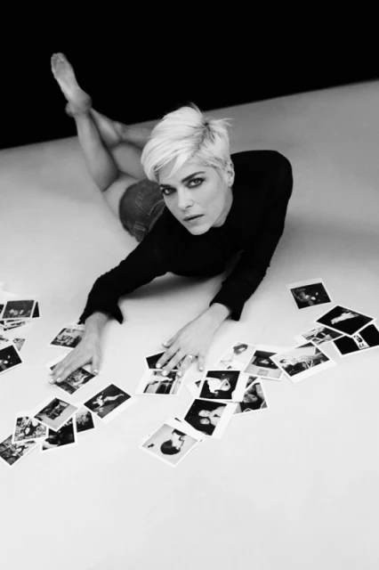 Black and white portrait of a blonde woman lying on the floor with scattered photographs, creating a visually artistic composition.