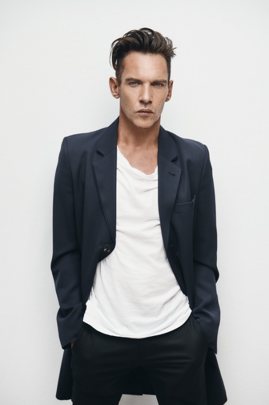 Portrait of a dark-haired adult man in a dark jacket and white t-shirt, posing in a photo studio with hands in his pockets.