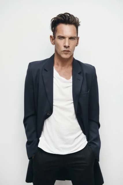 Portrait of a dark-haired adult man in a dark jacket and white t-shirt, posing in a photo studio with hands in his pockets.