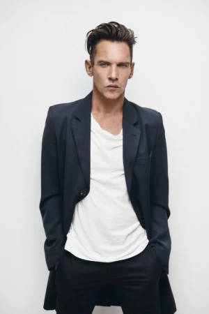 Portrait of a dark-haired adult man in a dark jacket and white t-shirt, posing in a photo studio with hands in his pockets.