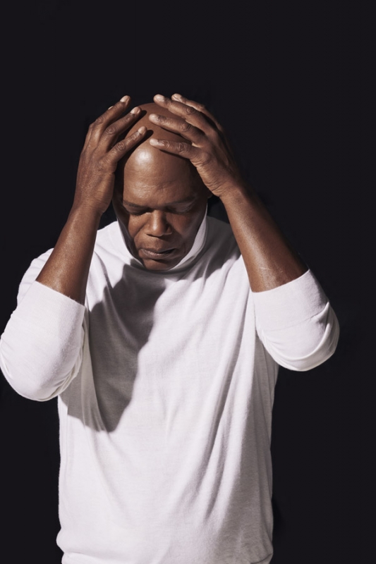 Portrait of an adult man with dark skin and a shaved head, wearing a white turtleneck, with his hands on his head against a dark background.
