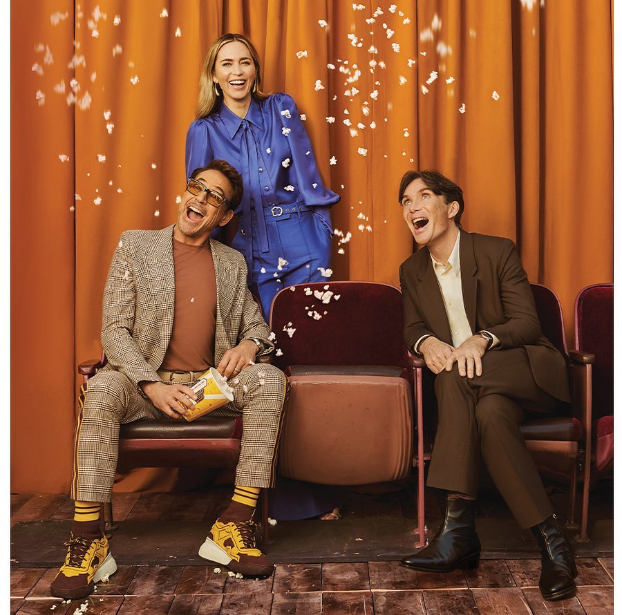 Three actors—two men and one woman—sit and stand in an indoor setting with falling popcorn, wearing suits and a blue jumpsuit, smiling and looking upward.