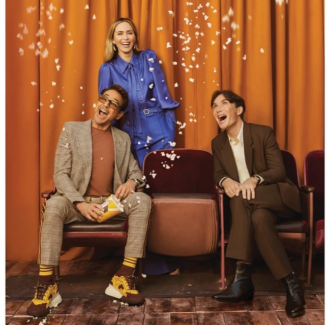 Three actors—two men and one woman—sit and stand in an indoor setting with falling popcorn, wearing suits and a blue jumpsuit, smiling and looking upward.