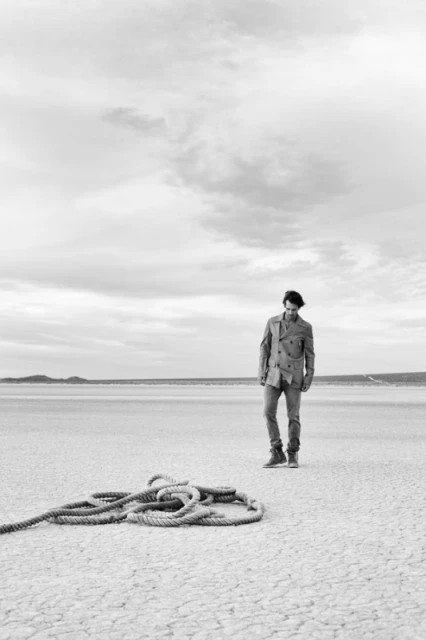 Monochrome shot of a man in a jacket and jeans standing on a cracked plane next to a coiled rope under an overcast sky.