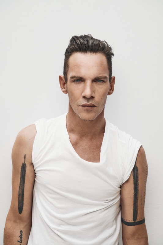 Portrait of a fair-skinned adult male with blue eyes wearing a sleeveless white top, showcasing arm tattoos against a white backdrop.