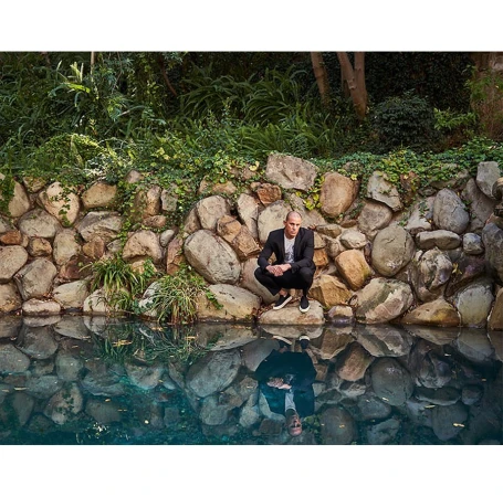 Man in a dark blazer crouching on stone wall by a pool reflecting his image, photographed by Brian Bowen Smith.