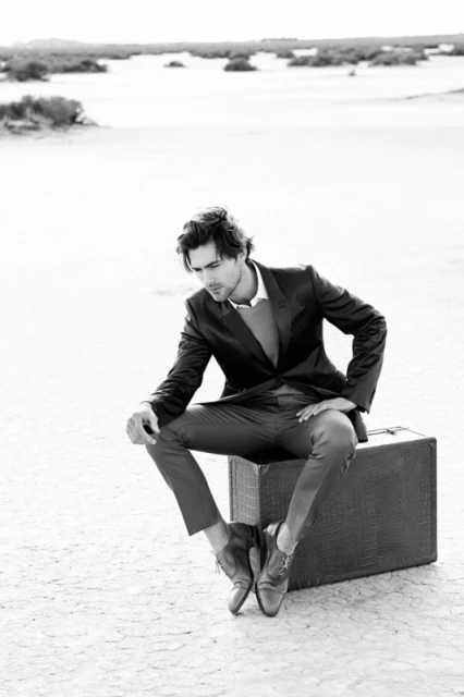 Elegant man in a suit sits on a suitcase in a desolate landscape. Dressed in tailored attire, he embodies sophistication in the stark setting.