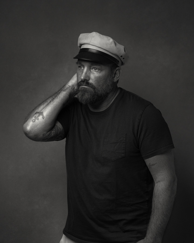 Monochrome portrait of a bearded man wearing a captain's hat, looking away with a hand behind his head in a photo studio setting.