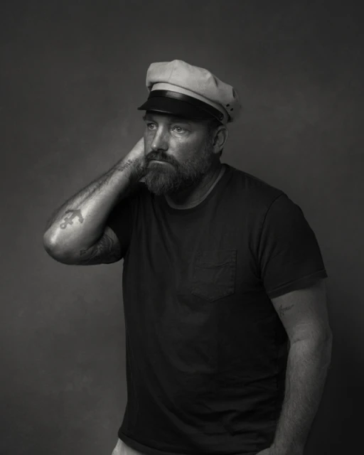 Monochrome portrait of a bearded man wearing a captain's hat, looking away with a hand behind his head in a photo studio setting.