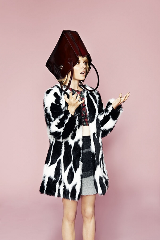 A fair-skinned woman in a fuzzy coat poses indoors with a brown leather bag over her head against a light pink background.