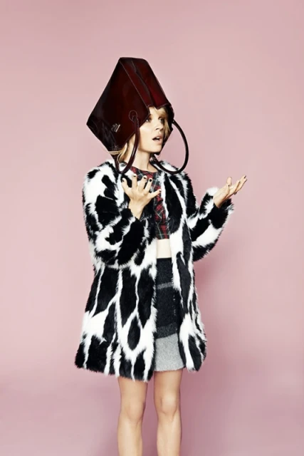 A fair-skinned woman in a fuzzy coat poses indoors with a brown leather bag over her head against a light pink background.