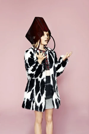 A fair-skinned woman in a fuzzy coat poses indoors with a brown leather bag over her head against a light pink background.