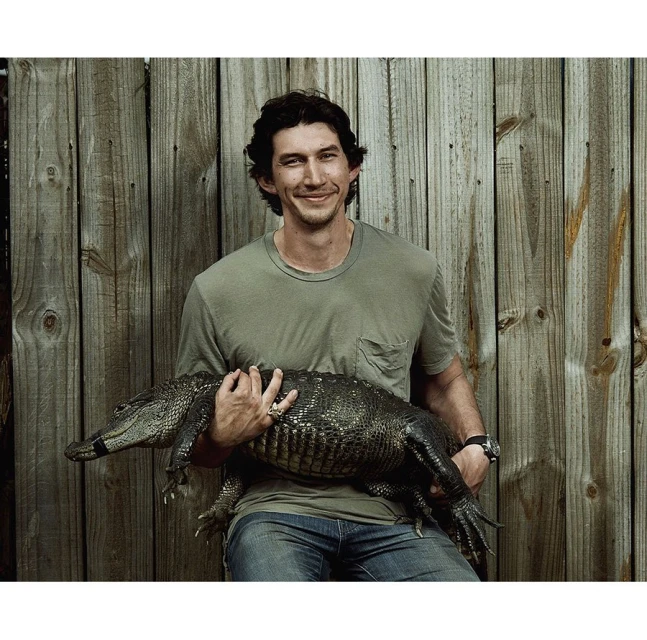 Adult male poses holding an alligator in front of a wood fence, wearing a t-shirt and jeans in an outdoor setting.